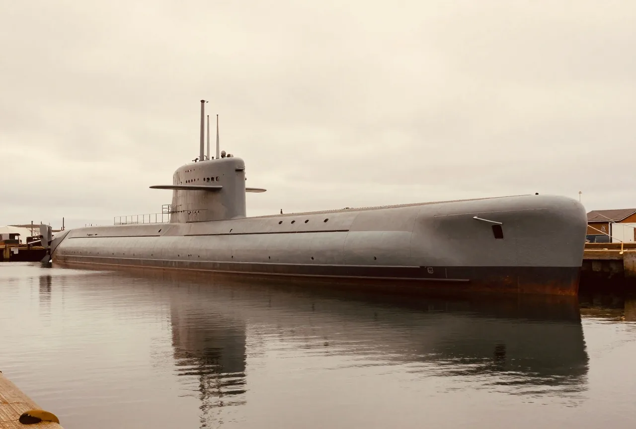 Ein langes graues historisches U-Boot liegt in einem ruhigen Hafenbecken, das Wasser spiegelt den Rumpf