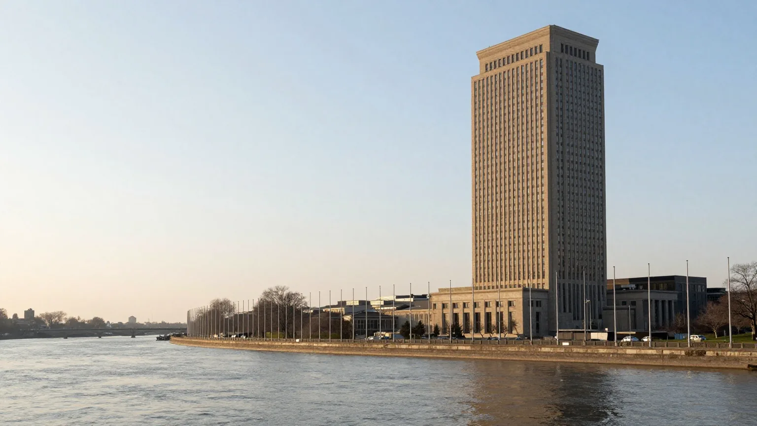 Ein schlankes Hochhaus aus Glas und Stein an einem breiten Fluss mit einer Reihe leerer Fahnenmasten im Morgenlicht