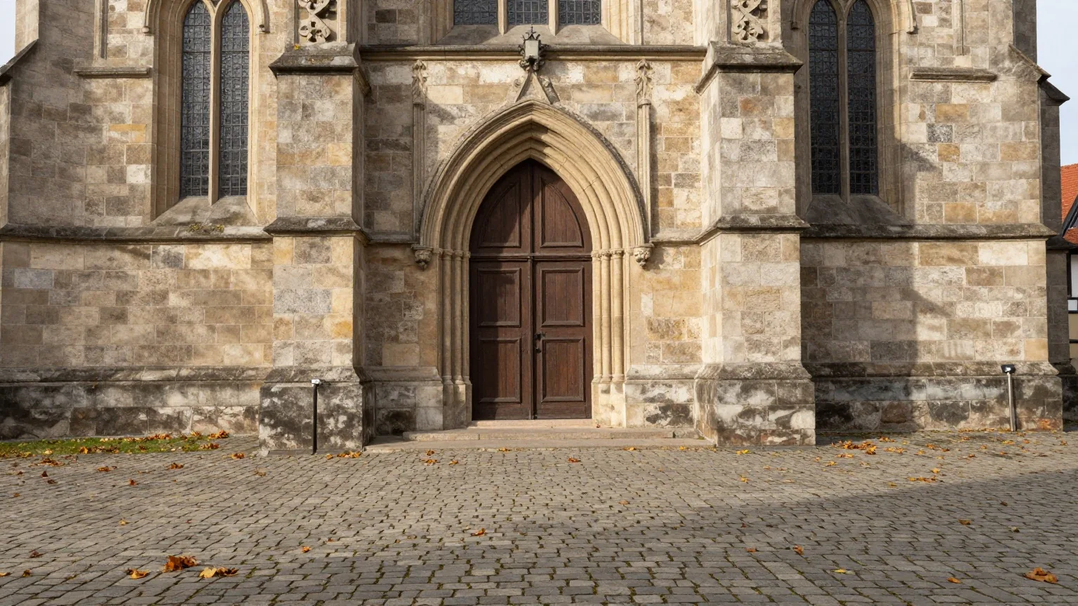 Die schlichte Holztür einer spätgotischen Steinkirche an einem leeren Platz mit Kopfsteinpflaster und Herbstlaub