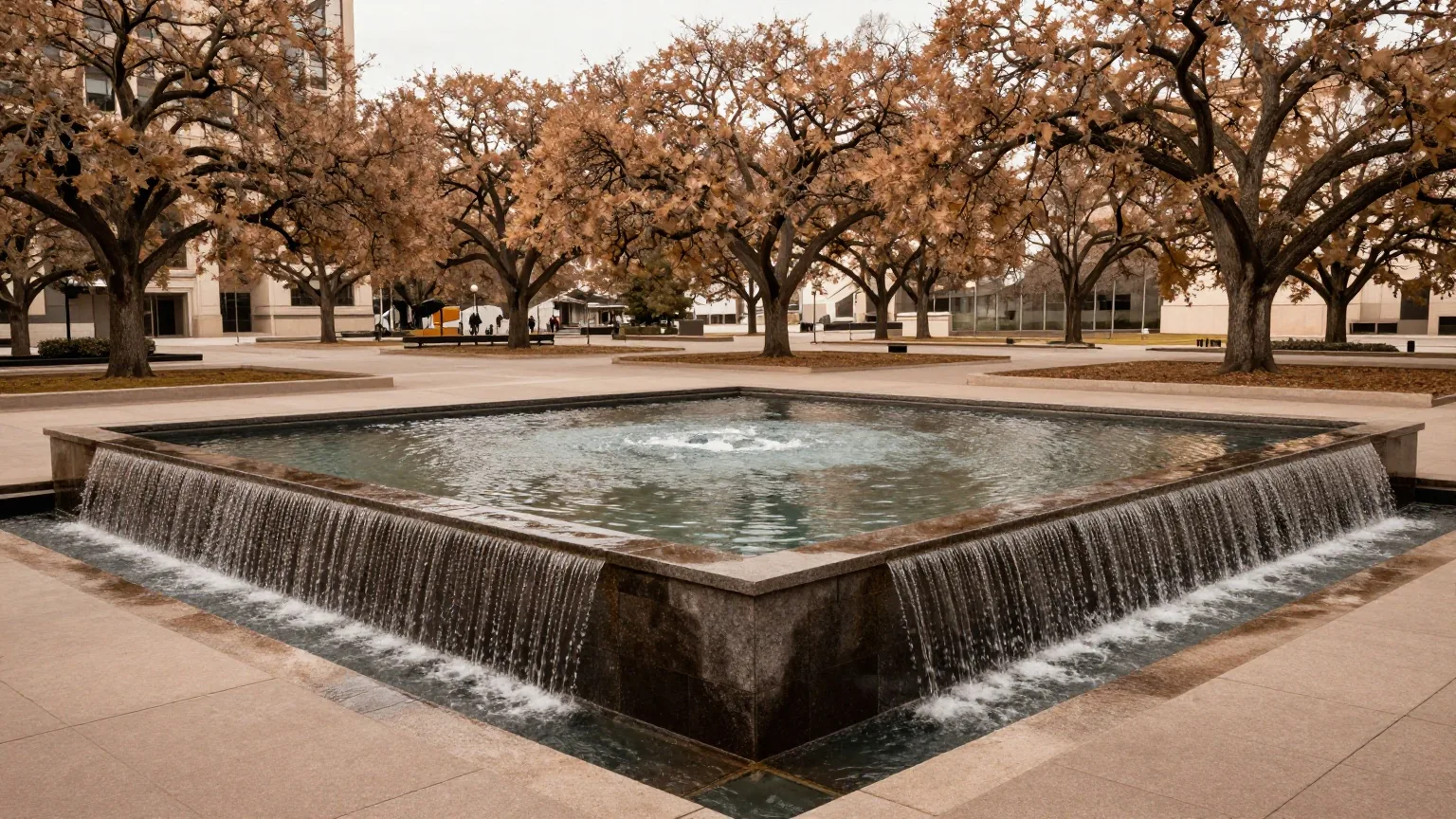 Zwei große quadratische Wasserbecken mit Wasserfällen an dunklen Steinwänden auf einem Platz mit Eichen im Morgenlicht