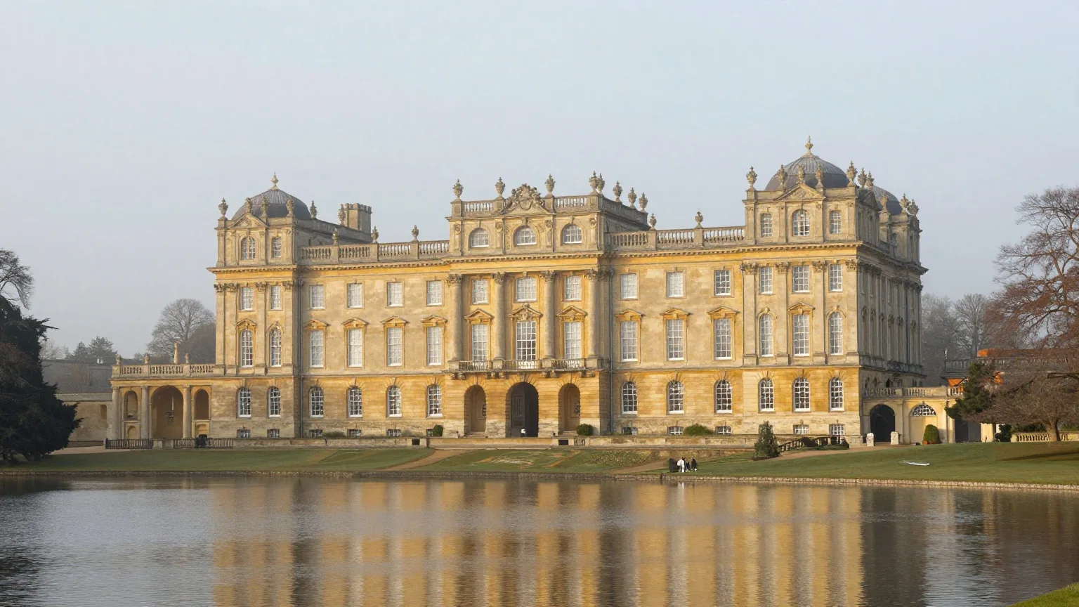 Ein großer barocker Palast aus honigfarbenem Stein hinter einem See und weiter Parklandschaft im Morgennebel